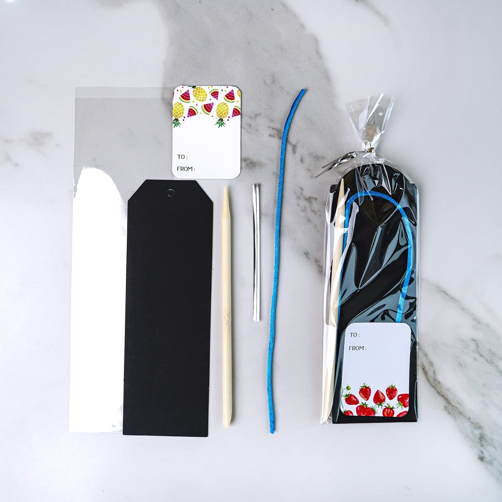 Scratch art bookmark craft kit contents including black and white bookmark blanks, wooden stylus, silver tool, blue cord, and gift tags with fruit designs.