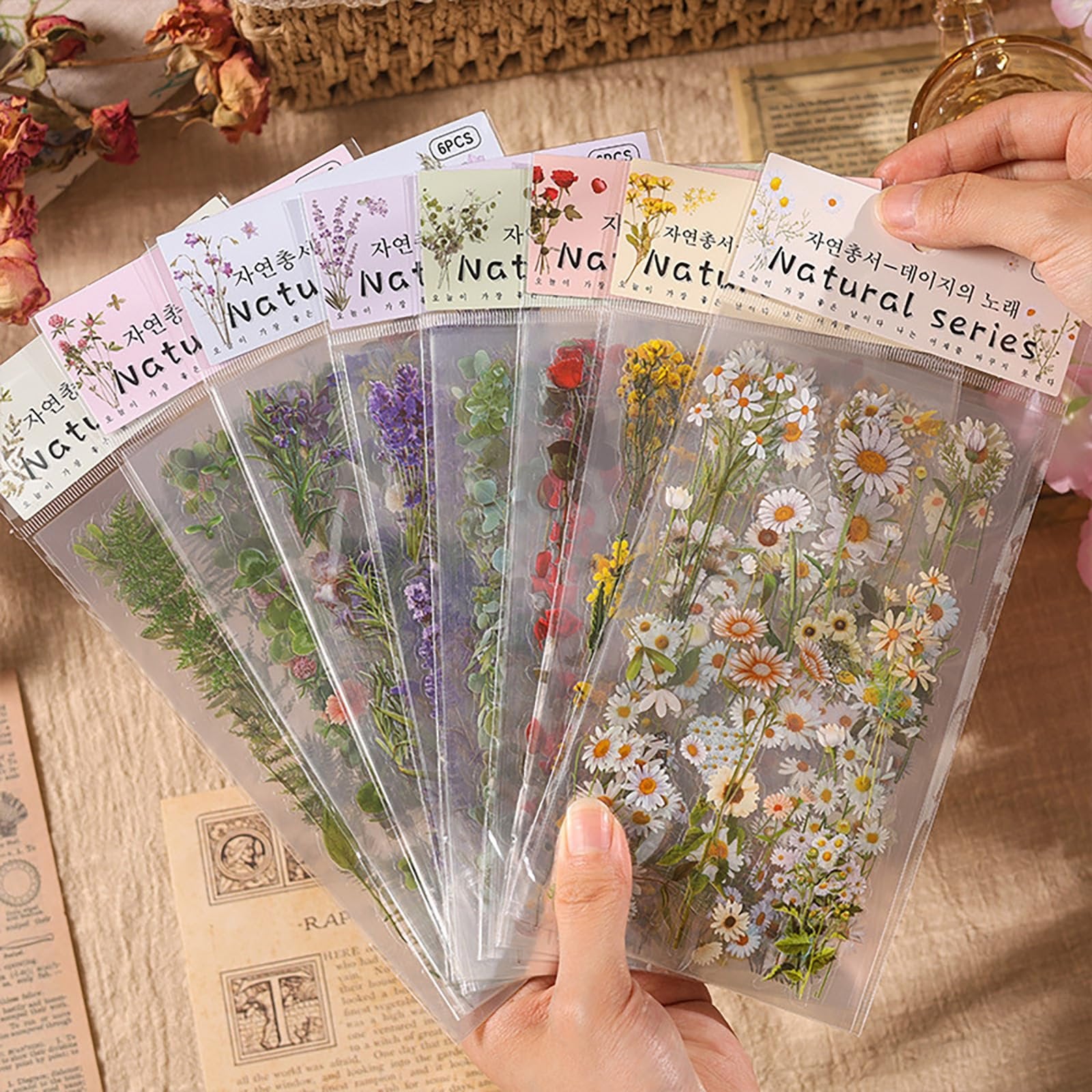 Fan of clear botanical sticker sheets featuring flowers and greenery, held by hands.