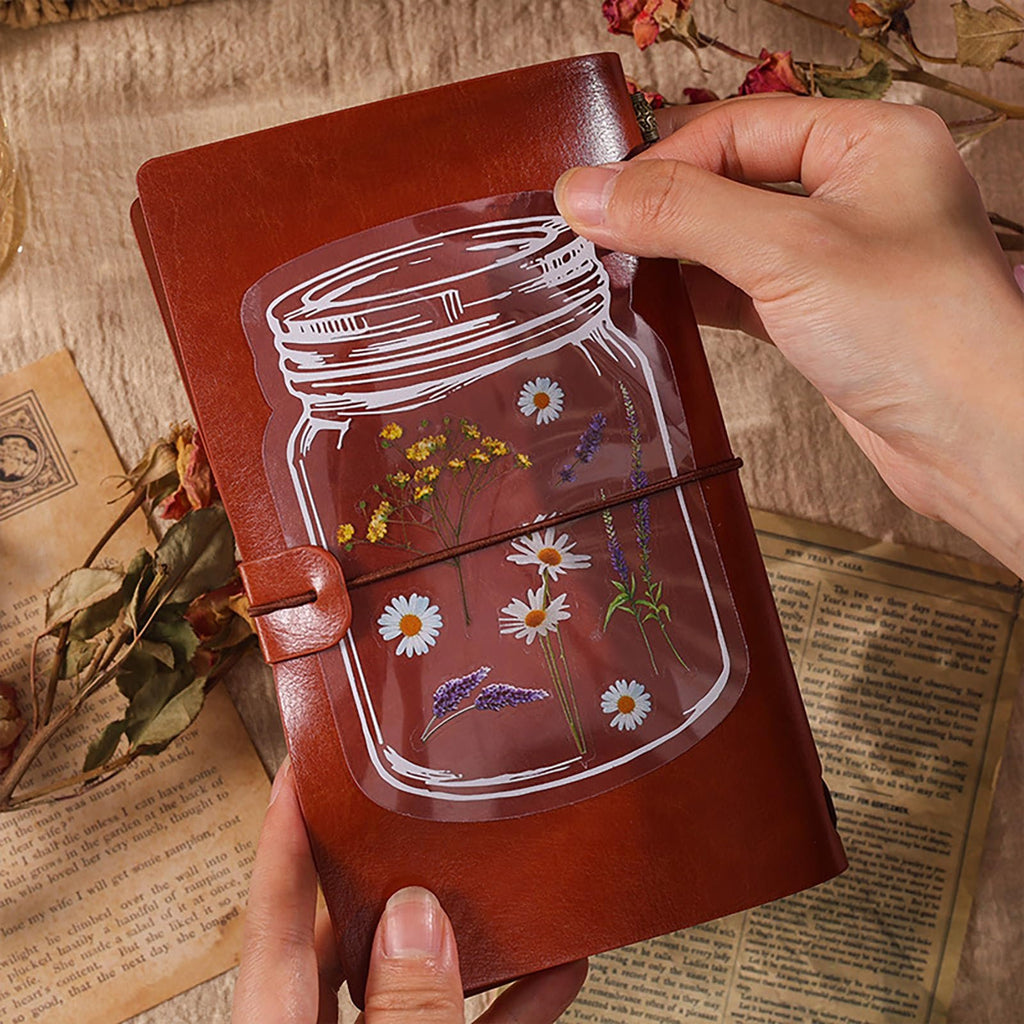 Clear sticker with a white outline of a mason jar filled with daisies and lavender, being applied to a brown journal.