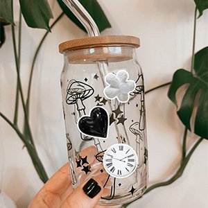 Hand holding a glass tumbler with a bamboo lid and straw, decorated with vintage aesthetic stickers of mushrooms, stars, a heart, a flower, and a clock.