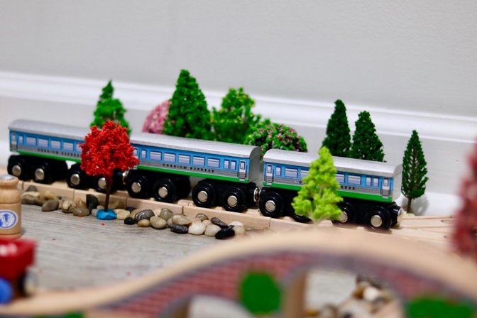 Wooden toy train cars with blue and silver livery on a track, surrounded by miniature trees and rocks.