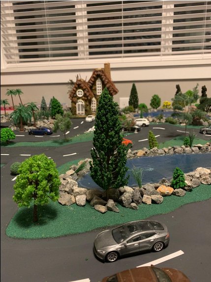 Diorama scene with model trees, a house, a pond, and toy cars on roads.