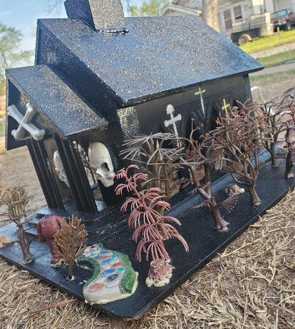 Black haunted house model with skulls, crosses, and miniature trees on a base, outdoors.
