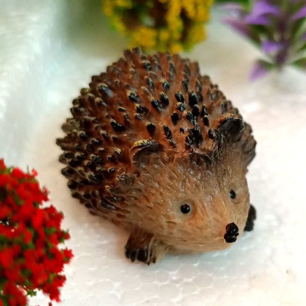 Plastic hedgehog figure with detailed spines, set against a white textured surface with blurred red, yellow, and purple flowers.