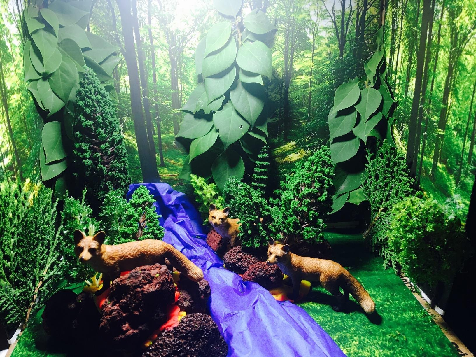 Diorama scene with three fox figures, model trees, bushes, and a blue stream.