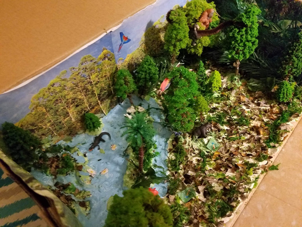 Diorama scene with miniature trees, bushes, animals, and a river, set inside a cardboard box.