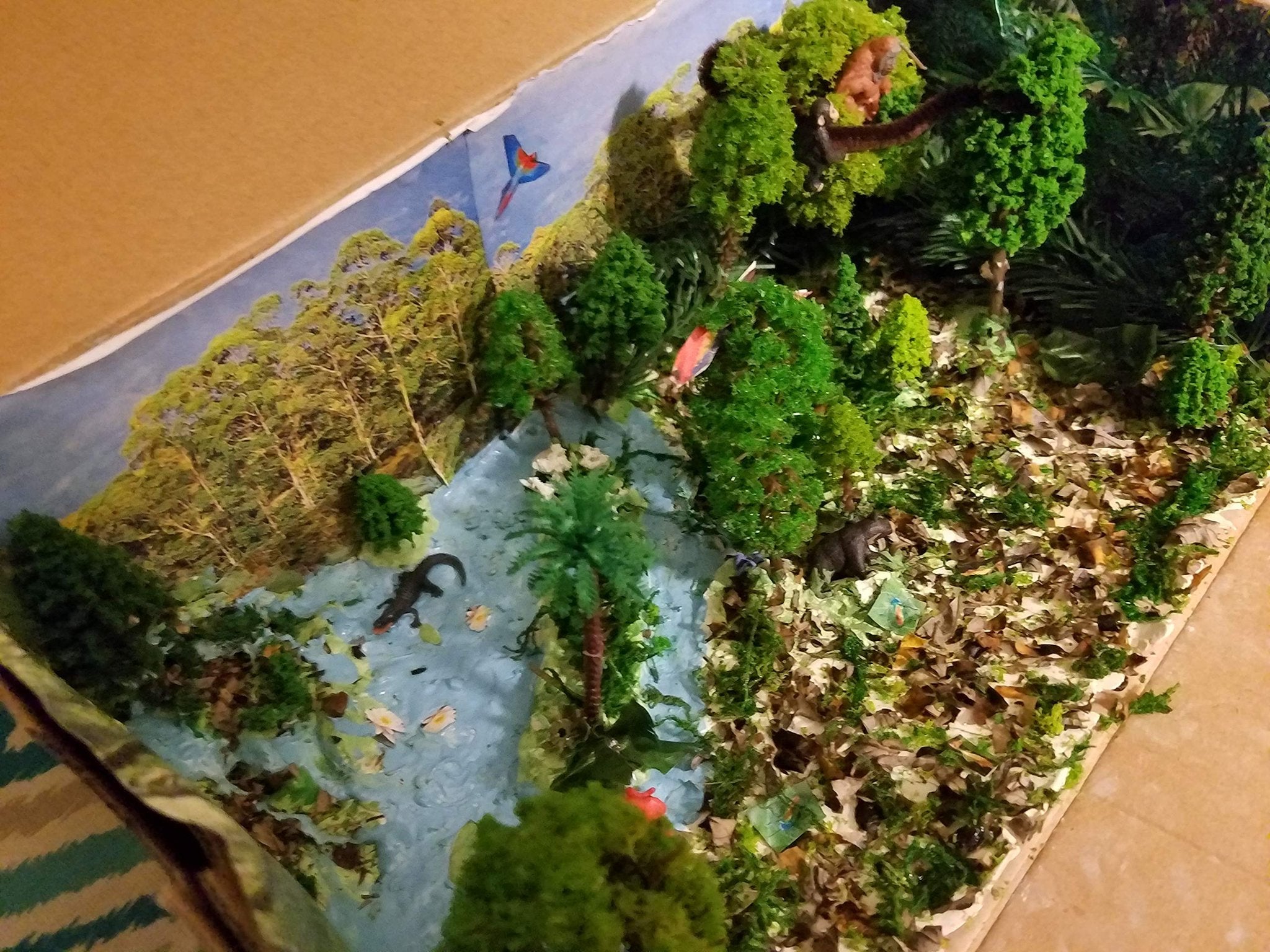 Diorama scene with miniature trees, bushes, animals, and a river, set inside a cardboard box.