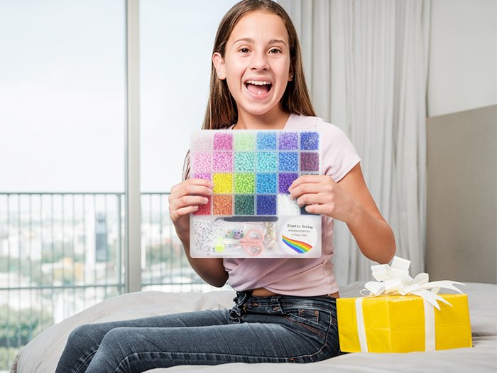 Excited girl holding a clear plastic case filled with colorful seed beads and jewelry making supplies, with a yellow gift box nearby.
