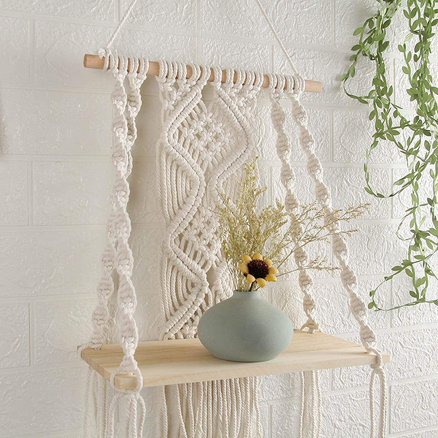 Macrame wall shelf with a vase and dried flowers, hanging on a white brick wall with green vines.