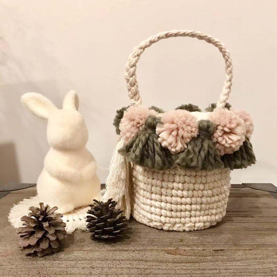 Crocheted basket with pink and green pom poms and tassels, next to a fluffy white bunny and pinecones.