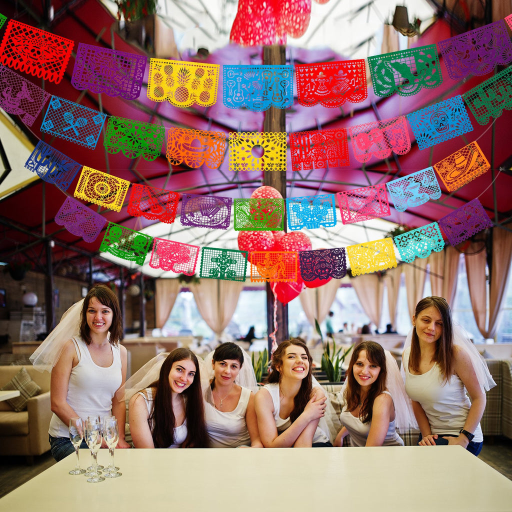 Group of six women wearing white tank tops and veils, seated at a table with champagne glasses, under colorful papel picado banners.