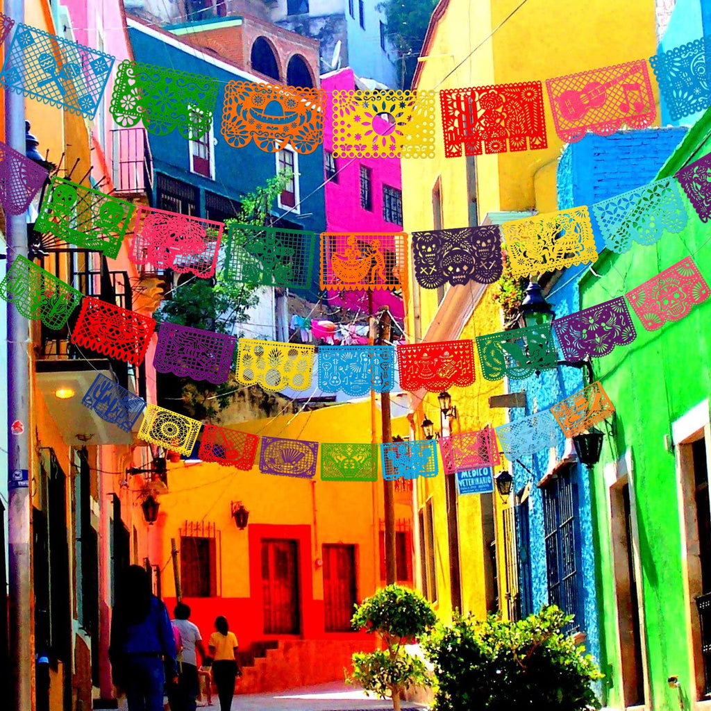 Colorful Papel Picado banners strung across a vibrant street with brightly painted buildings and people walking.