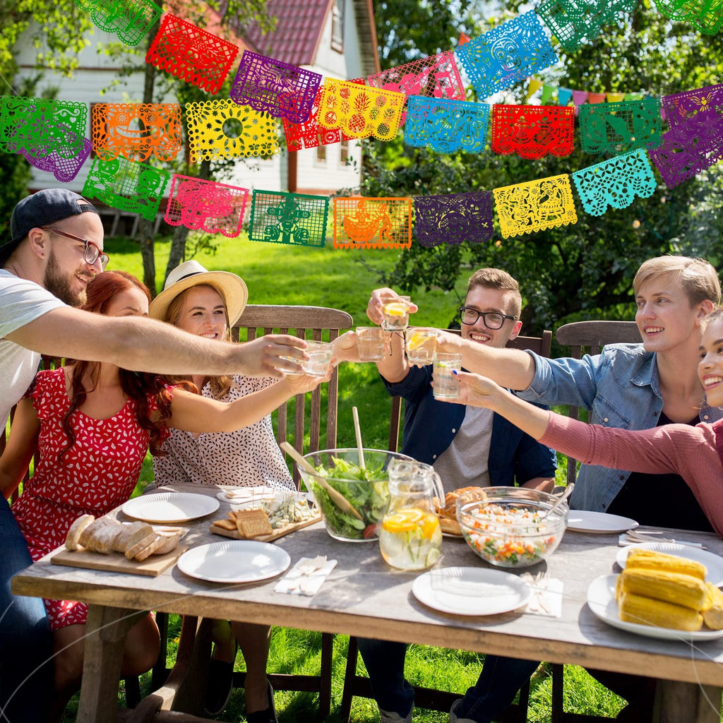 People toasting drinks at a table outdoors with colorful papel picado banners overhead