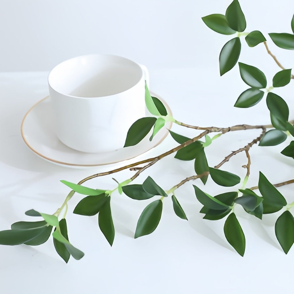 Artificial ficus twig branch with green leaves next to a white teacup and saucer