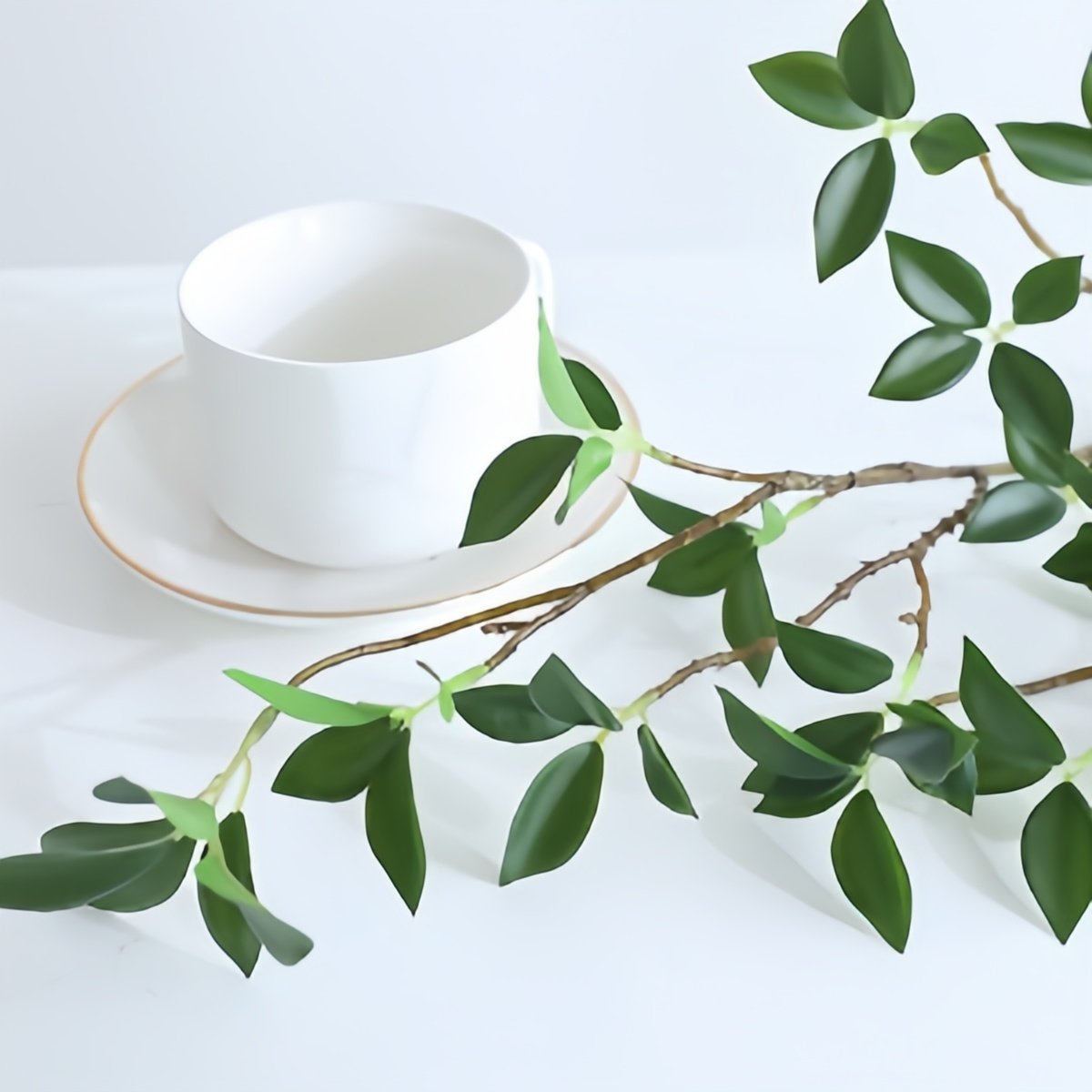 Artificial ficus twig branch with green leaves next to a white teacup and saucer