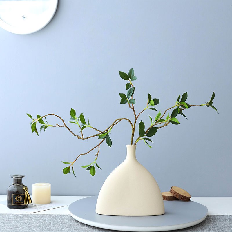 Faux ficus twig branches with green leaves in a cream vase on a gray tray with decorative items.