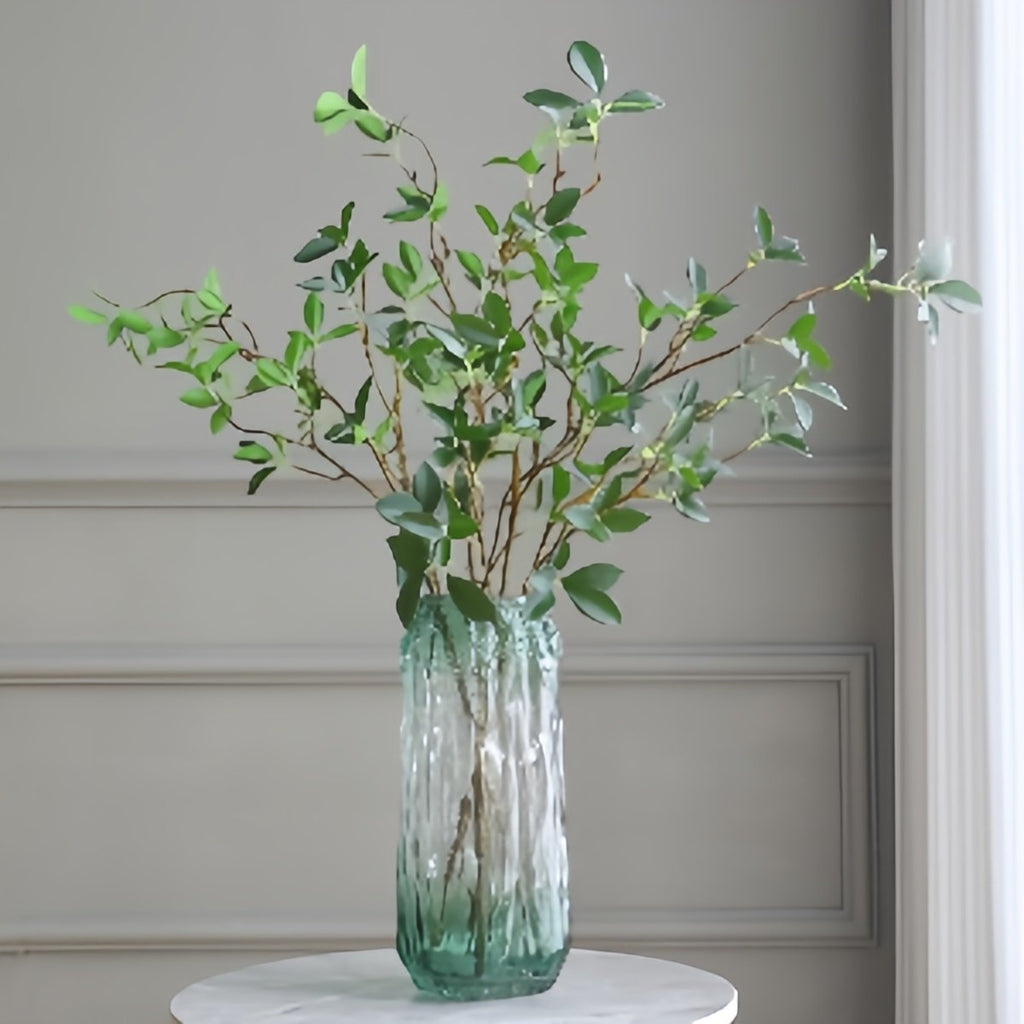 Faux ficus twig branches with green leaves in a textured glass vase on a marble table.