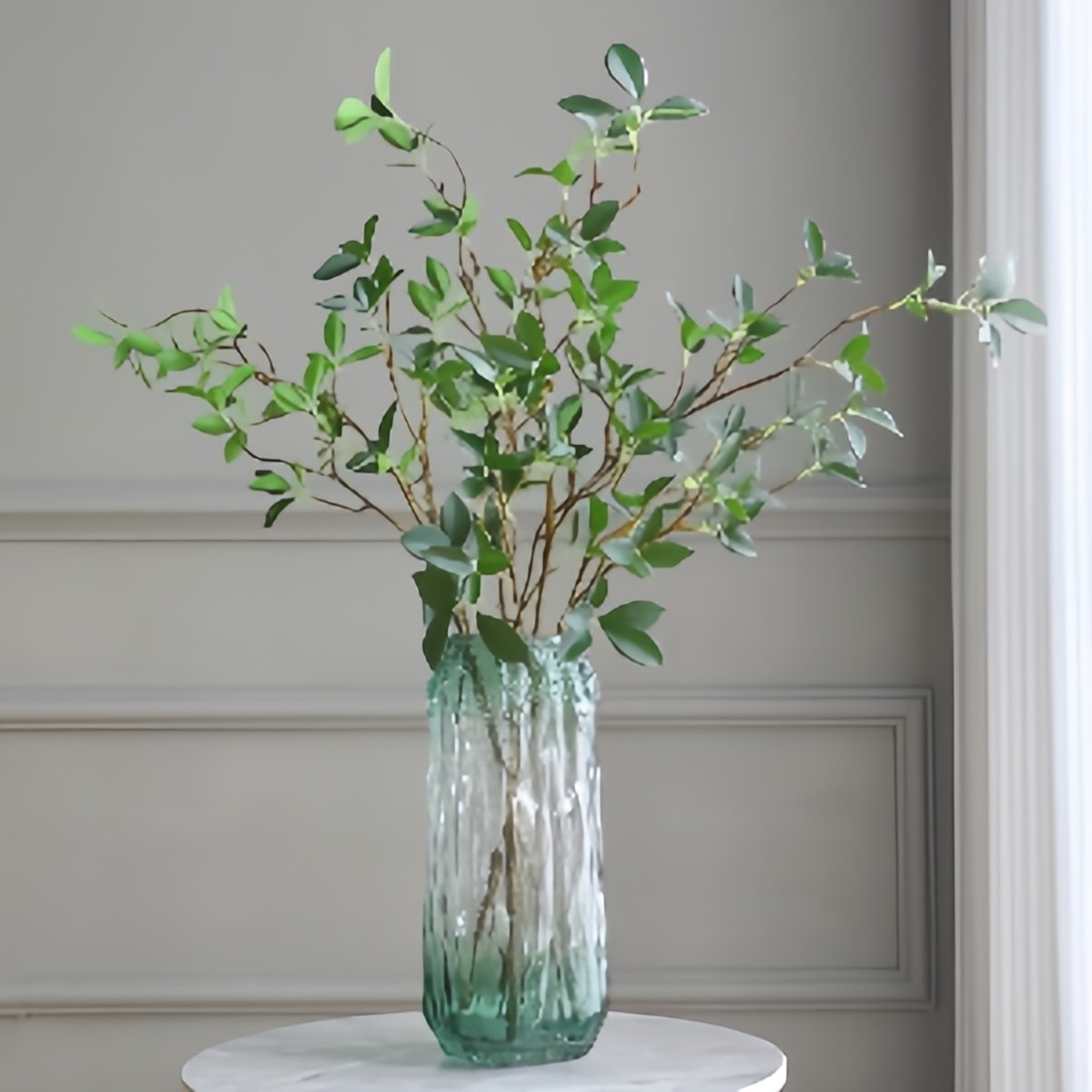 Faux ficus twig branches with green leaves in a textured glass vase on a marble table.