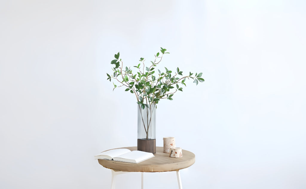 Faux ficus twig branches in a glass vase on a wooden table with an open book and decorative wood pieces.