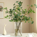 Faux ficus twig branches with green leaves in a clear ribbed vase on a table.