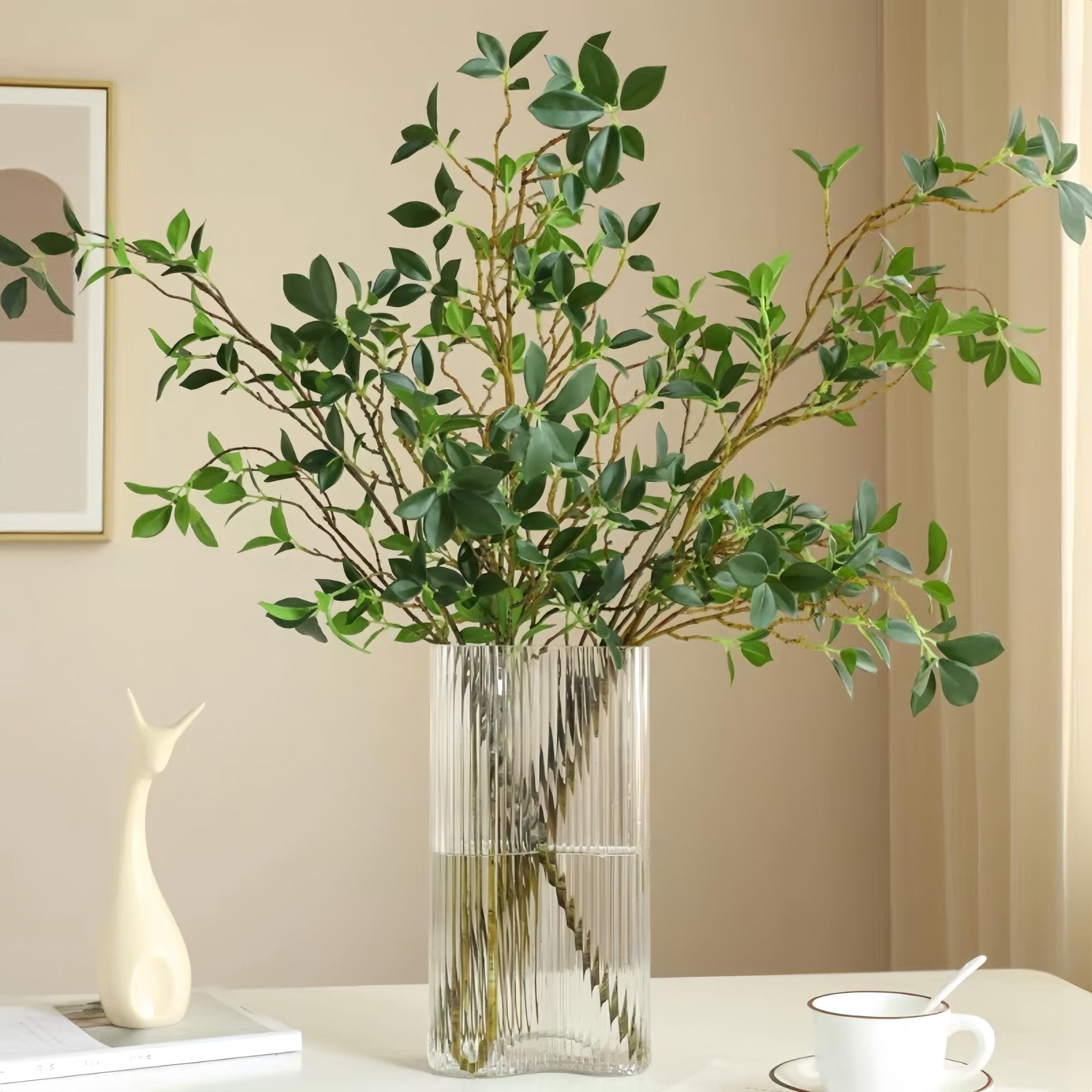 Faux ficus twig branches with green leaves in a clear ribbed vase on a table.