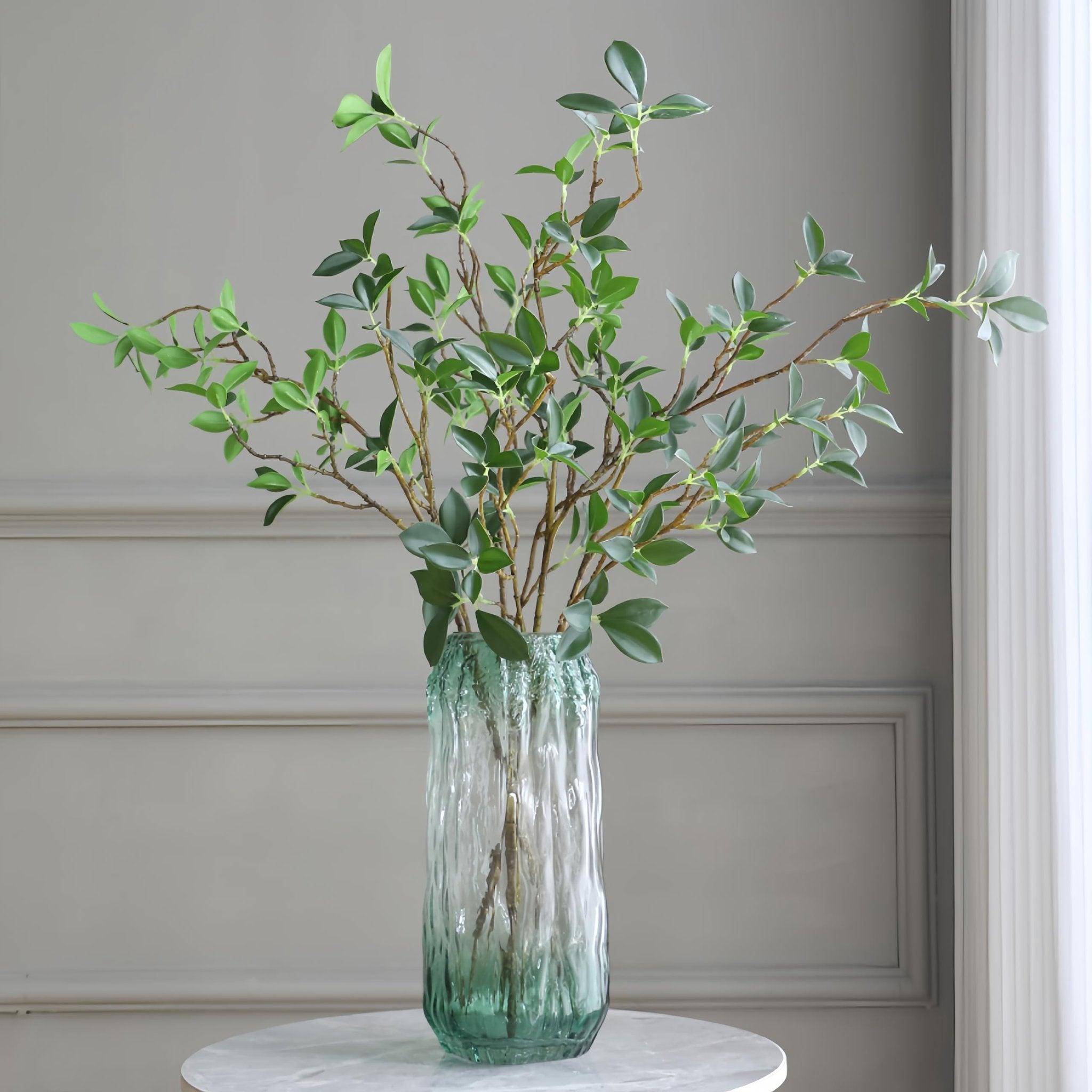Four faux ficus twig branches with green leaves in a textured green glass vase on a marble table.