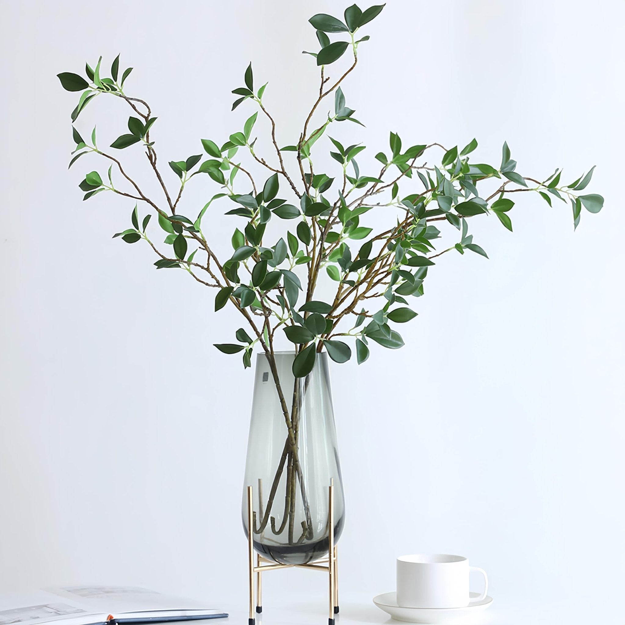 Faux ficus twig branches with green leaves in a grey vase on a stand, with a book and cup nearby.