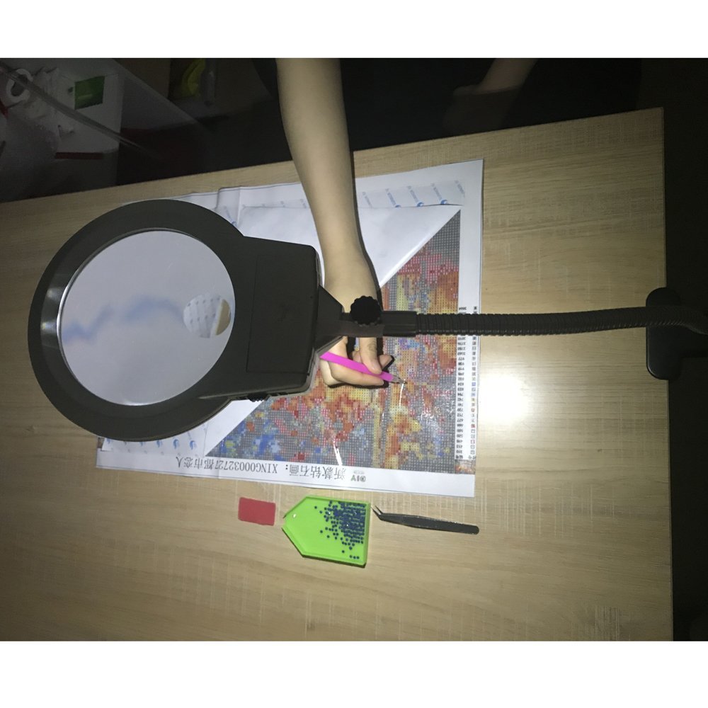 Diamond painting magnifier lamp with adjustable arm over a diamond painting kit, held by a person's hand.