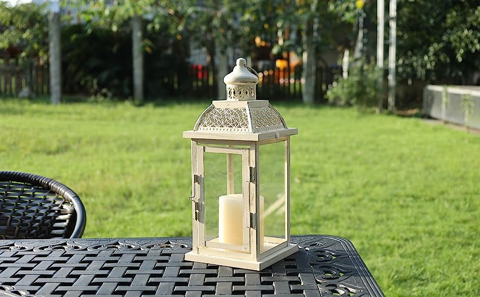 Cream metal and glass candle lantern with a cream candle inside, placed on a black metal table outdoors.