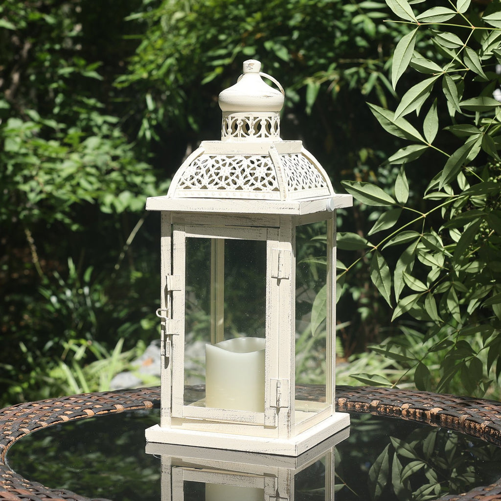 White metal and glass candle lantern with a cream LED candle inside, set on a reflective table outdoors.