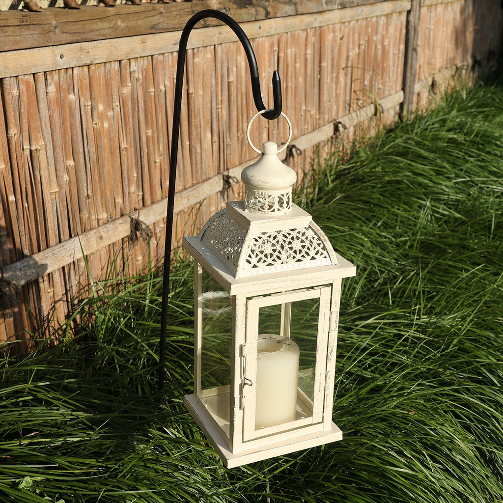 White metal and glass candle lantern with a cream candle inside, hanging from a black hook in front of a bamboo fence and green grass.