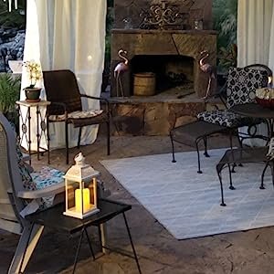 White metal and glass candle lantern with lit candle inside, placed on a small black table next to a chair on a patio.