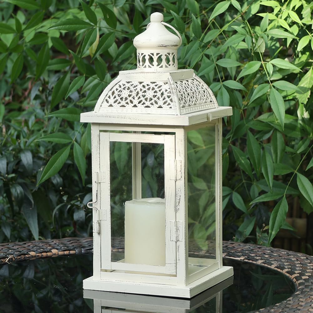White metal and glass candle lantern with a cream candle inside, set on a glass table with greenery in the background.