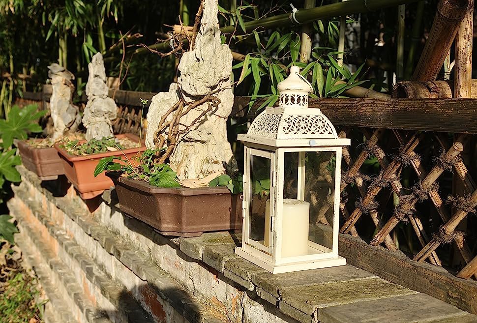 White metal and glass candle lantern with a cream candle inside, placed on a stone ledge with potted plants and bamboo in the background.