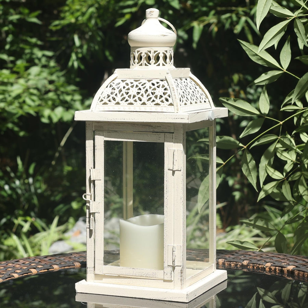 White metal and glass candle lantern with decorative filigree top, placed on a glass table outdoors.