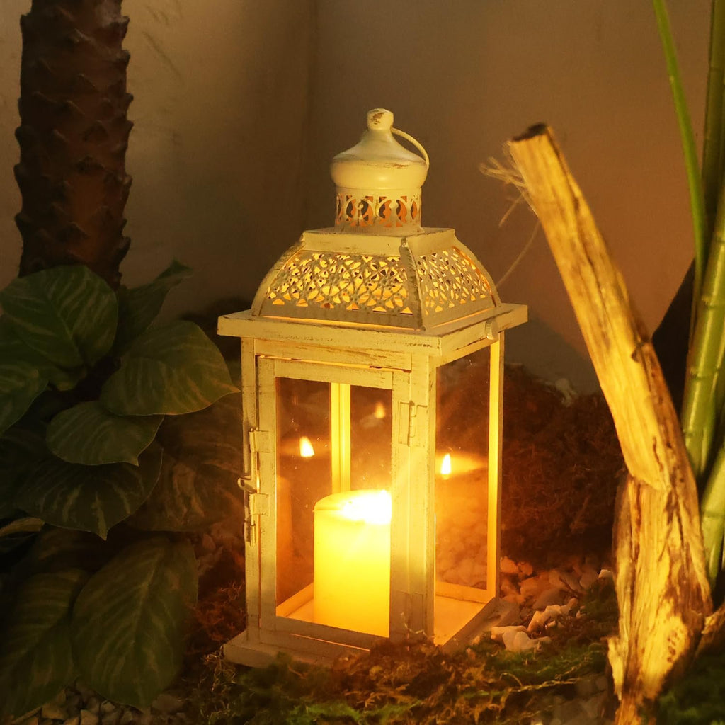 White metal and glass candle lantern with a lit candle inside, set amongst plants and wood.