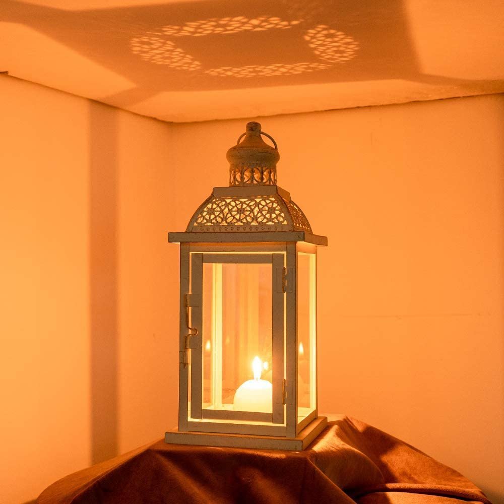 White metal and glass candle lantern with a lit candle inside, casting decorative shadows on the wall.