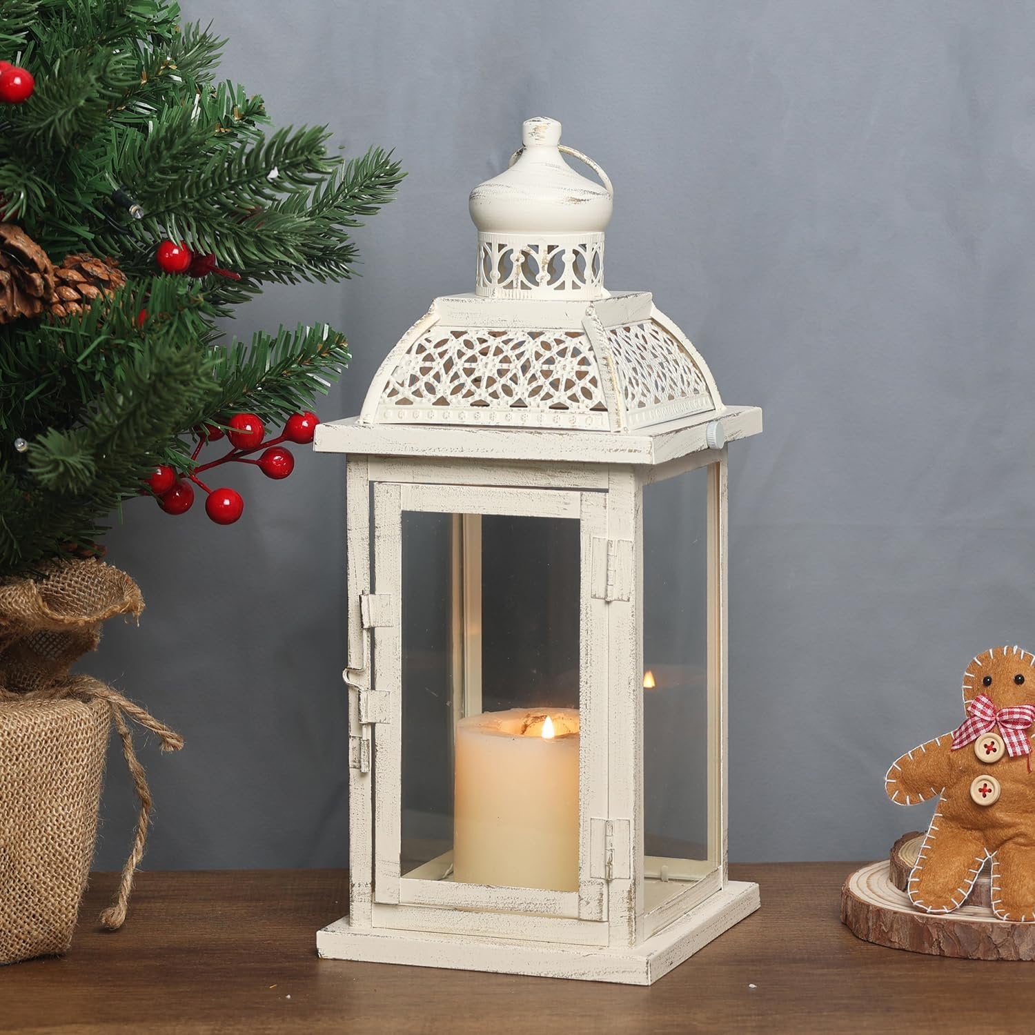 White metal and glass candle lantern with a lit candle inside, next to a Christmas tree and gingerbread man decoration.