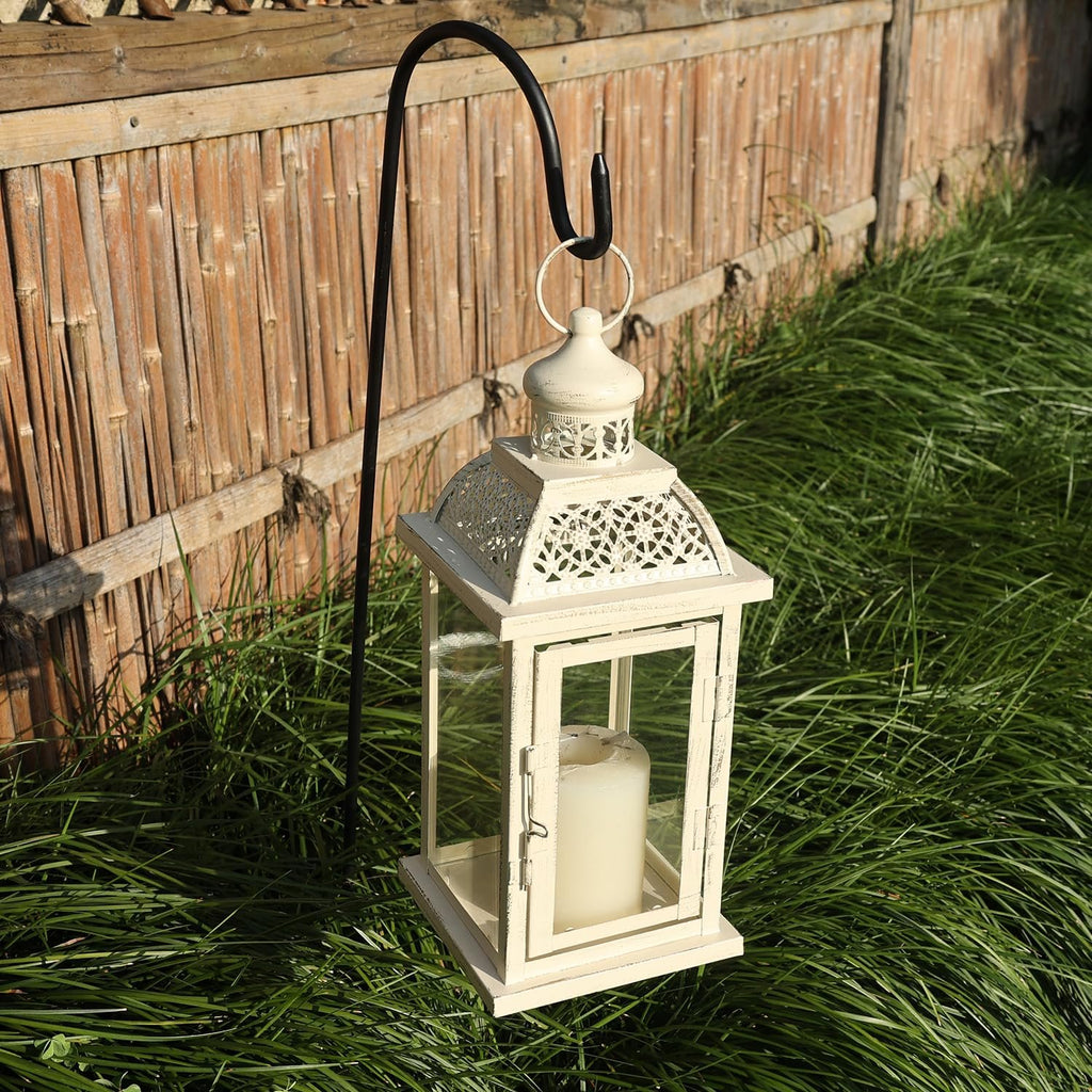 White metal and glass candle lantern with a cream candle inside, hanging from a black shepherd's hook in front of a bamboo fence and green grass.
