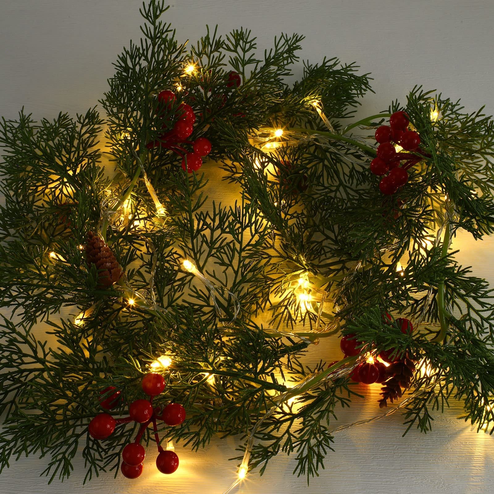 Close-up of a prelit Christmas garland with warm white LED lights, pine needles, pine cones, and red berries.