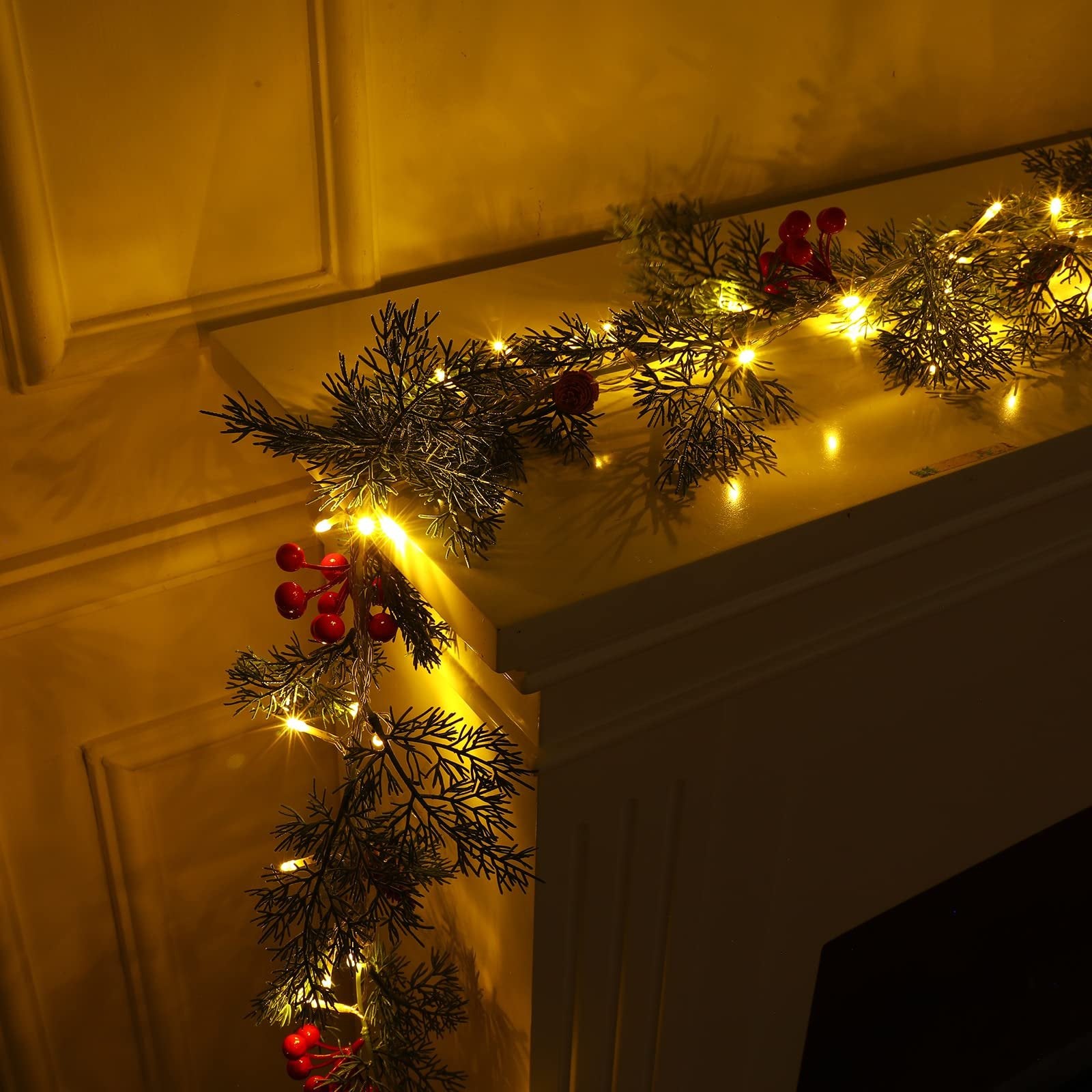 Prelit Christmas garland with warm white LEDs, pine needles, red berries, and pine cones draped over a mantelpiece.