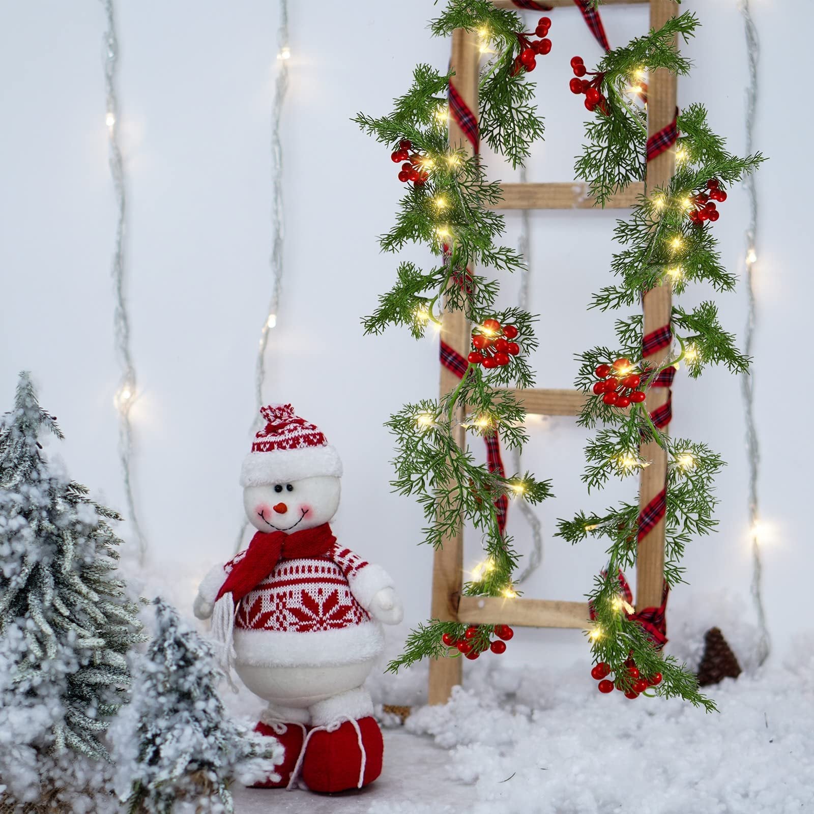 Christmas garland with warm white LEDs, pine needles, red berries, and plaid ribbon draped on a wooden ladder, with a snowman and snowy trees.