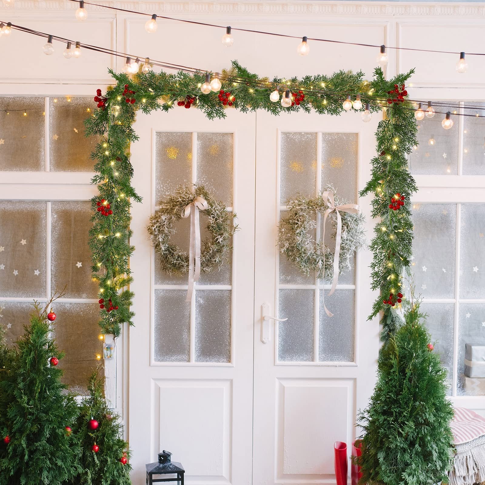 Christmas garland with warm white LEDs, pine needles, red berries, and pine cones draped over a white door with two wreaths