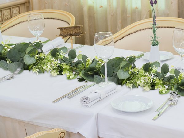 Artificial eucalyptus garland with white blooms draped across a table set for a meal, featuring wine glasses and cutlery.