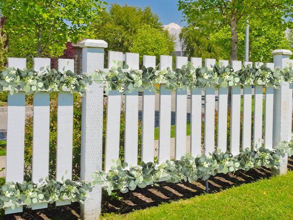 Artificial eucalyptus garland with white blooms draped over a white picket fence outdoors.