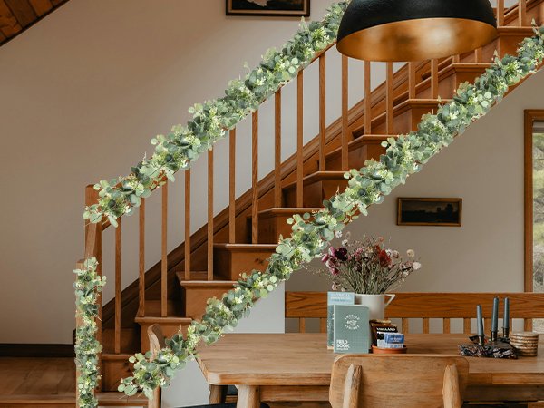 Artificial eucalyptus garland with white blooms draped along a wooden staircase railing and table