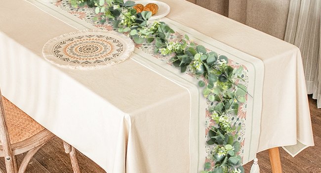 Artificial eucalyptus garland with white blooms draped across a table runner on a cream tablecloth.