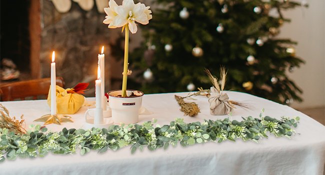 Artificial eucalyptus garland with white blooms draped across a table with candles and a Christmas tree in the background.
