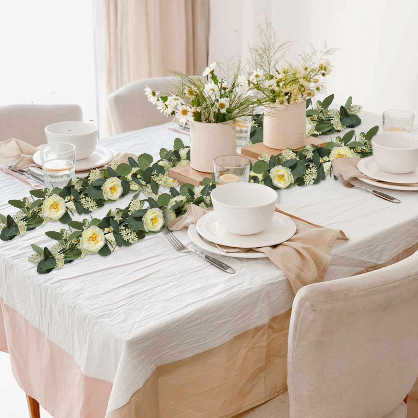 Artificial eucalyptus garland with white roses draped across a dining table setting with place settings and floral centerpieces.