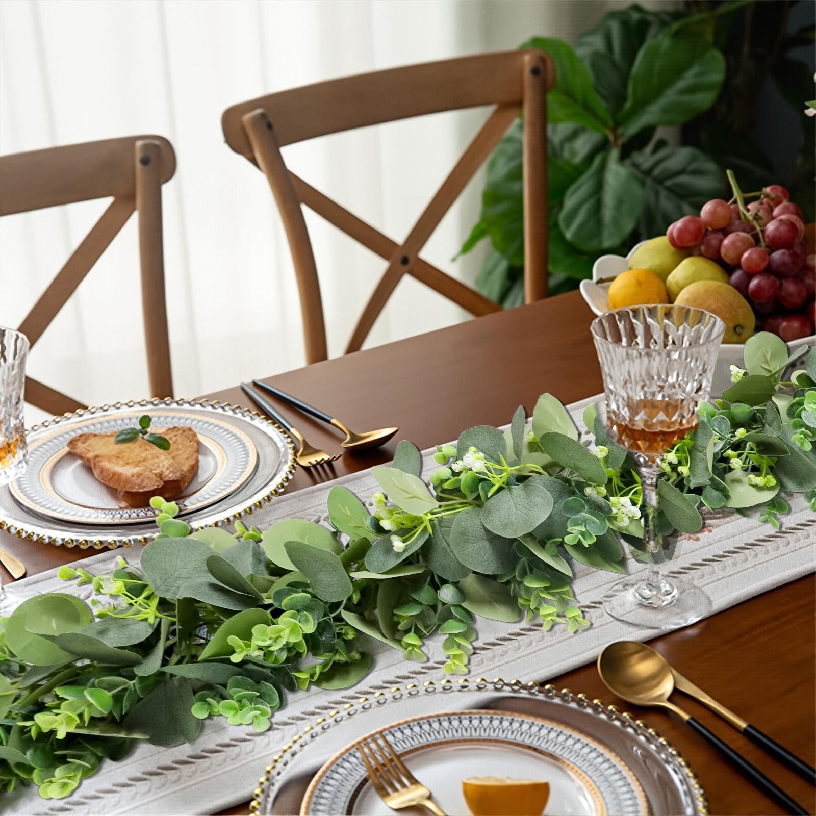 Artificial eucalyptus garland with white blooms draped across a dining table setting with place settings and fruit bowl.